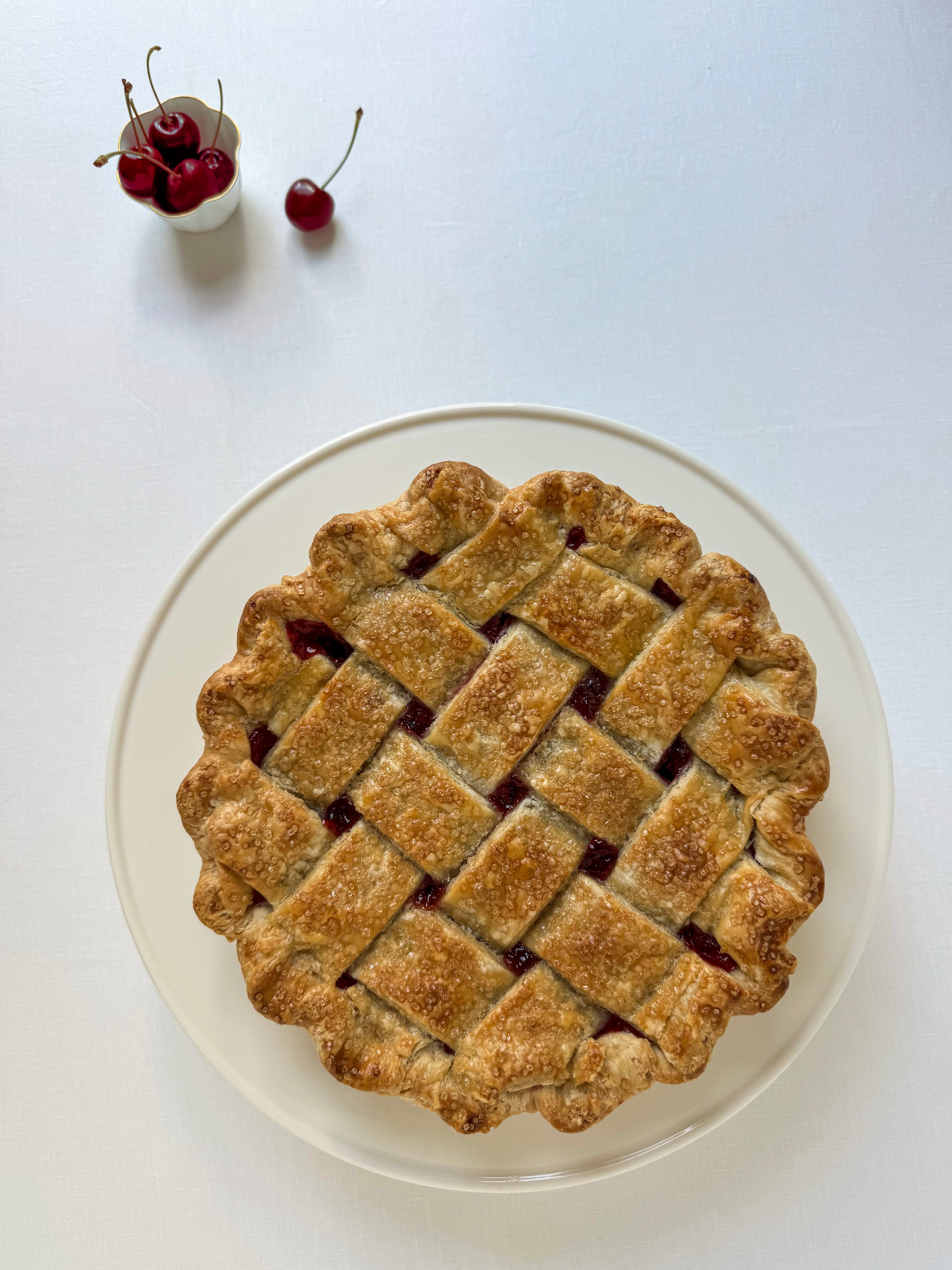 Cherry pie with lattice crust on a white plate, small bowl of cherries in the corner.