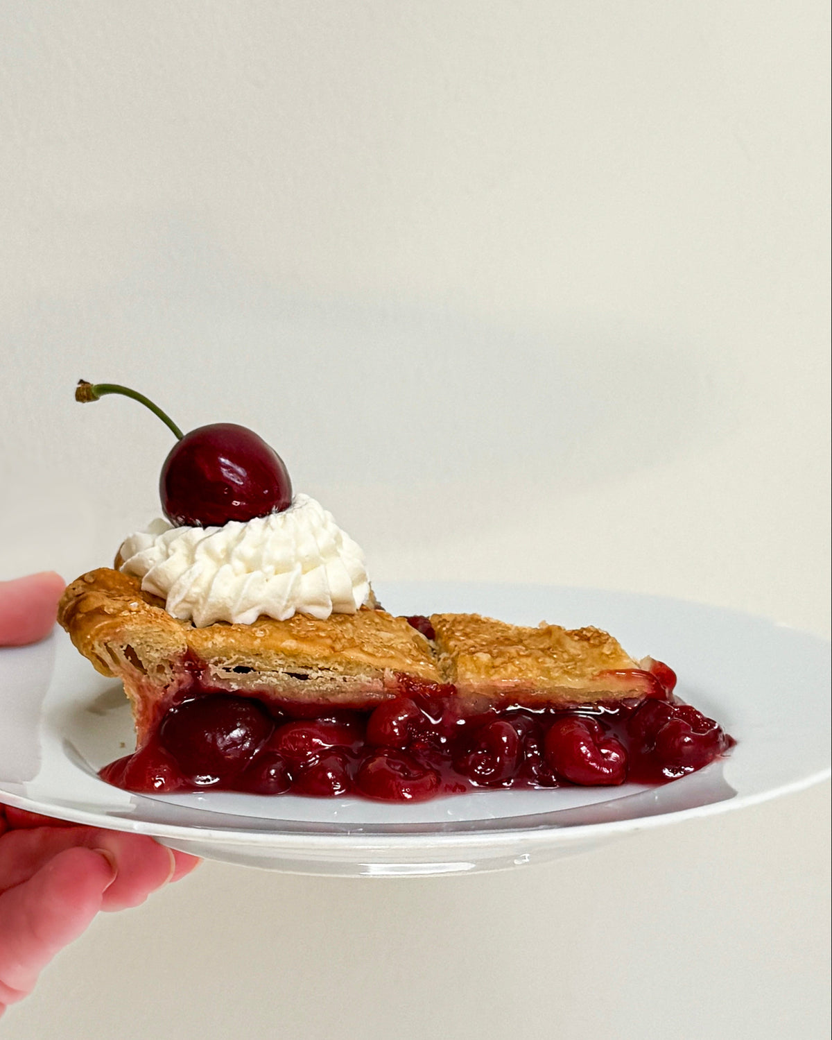 Slice of cherry pie with whipped cream and a cherry on top, held on a white plate.